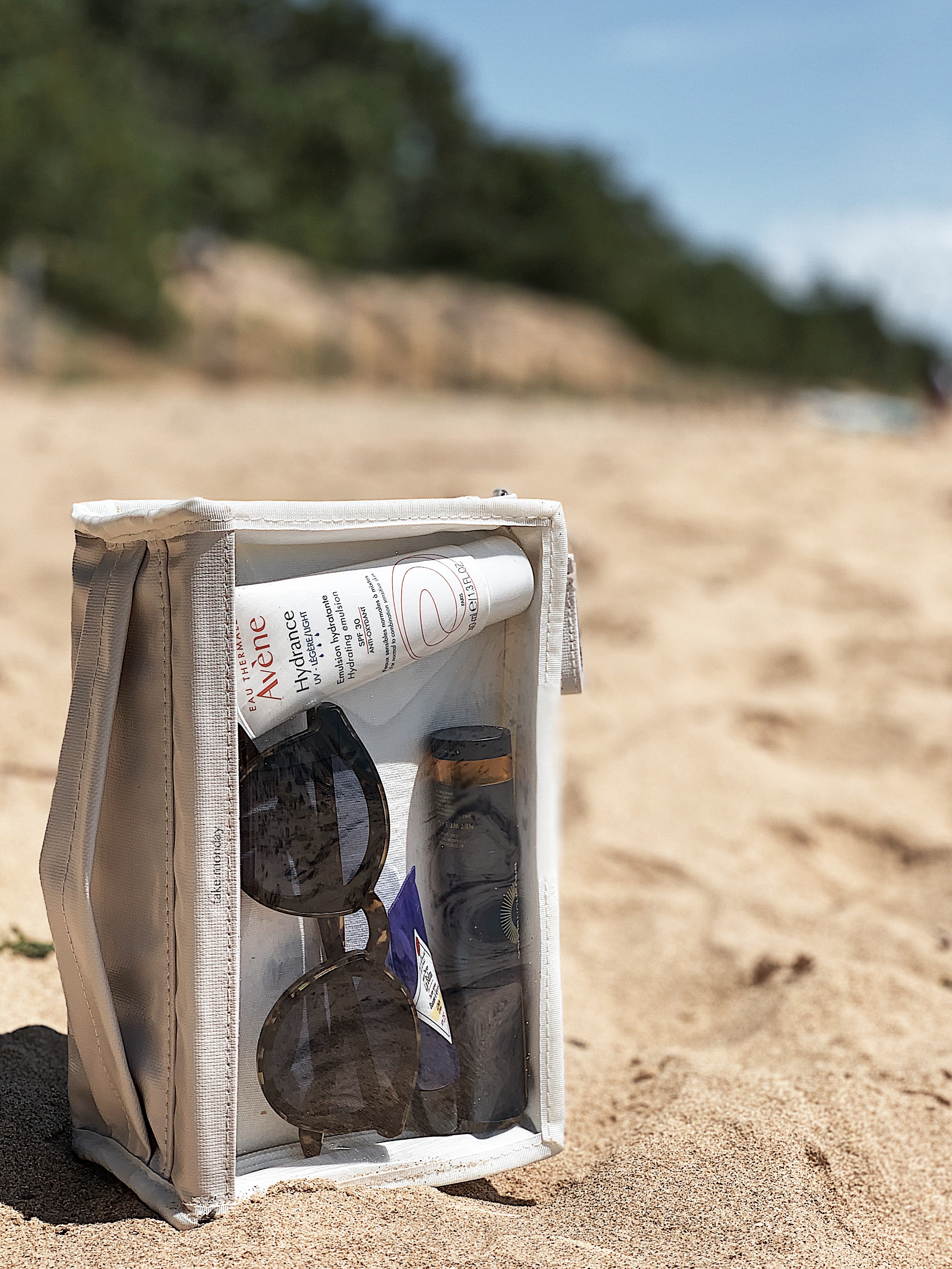 Photography of a beige 'take monday' travel pouch on a sunny sandy beach holding an Avène Hydrance UV Light Hydrating Emulsion SPF30 tube, dark sunglasses, and a small dark product bottle against blurred green foliage and blue sky