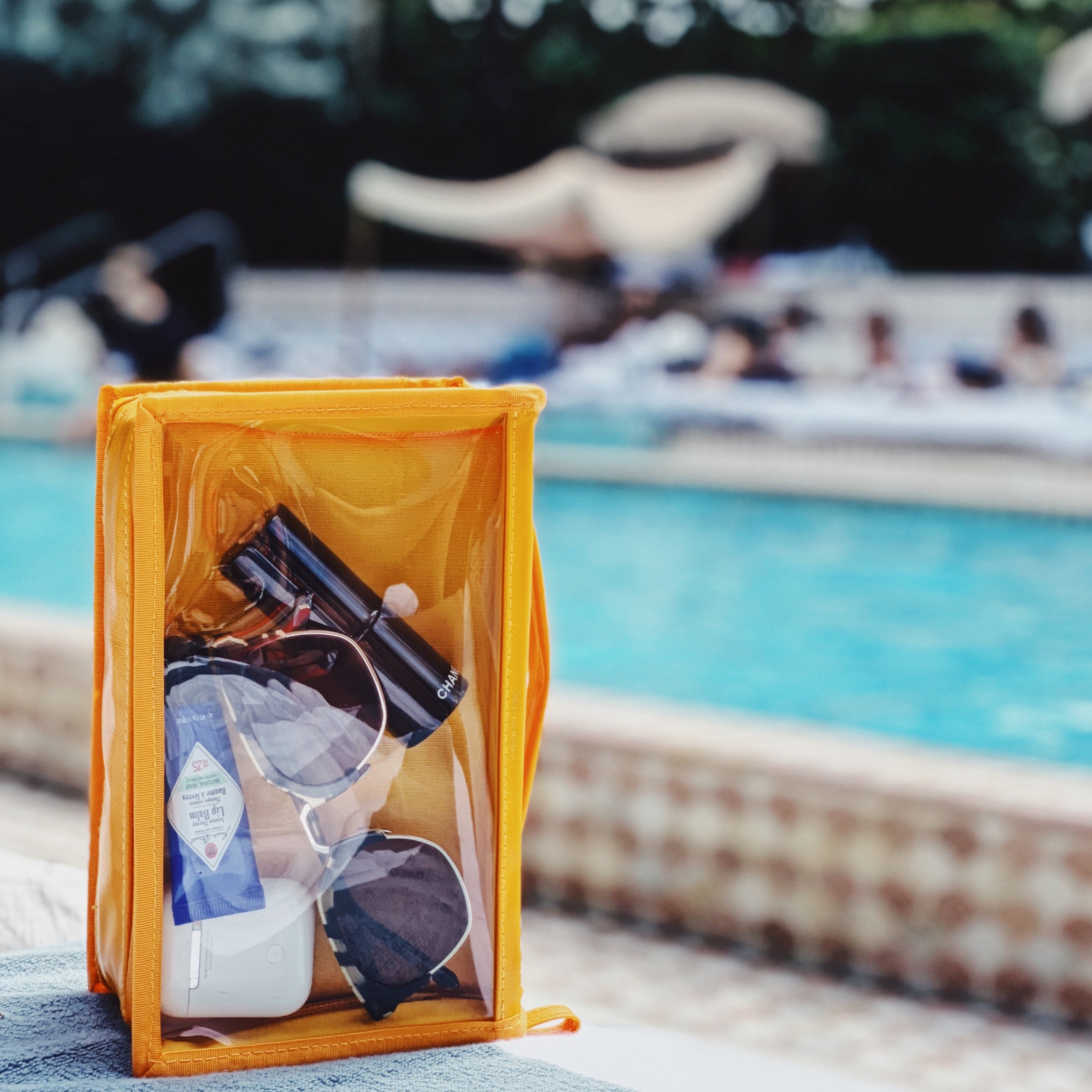 A yellow transparent pouch filled with Chanel mascara, sunglasses, Dr Bronner's lip balm, and an AirPods case rests on a towel beside a vibrant blue swimming pool with a decorative tiled edge