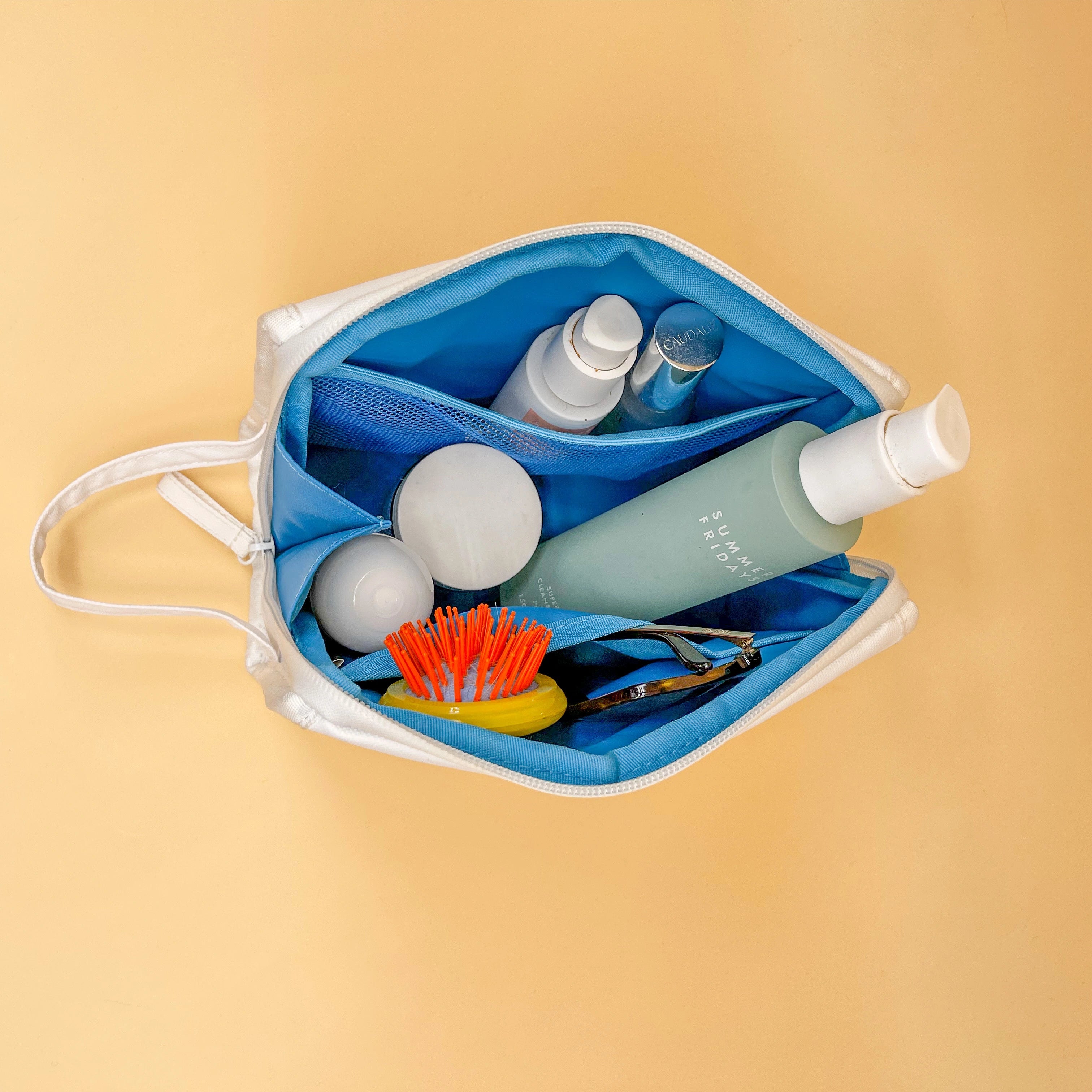 Photograph of an open white and blue cosmetic bag on a peach background containing a Caudalie bottle, a light blue Summer Fridays bottle, a bright orange and yellow hairbrush, and brown sunglasses