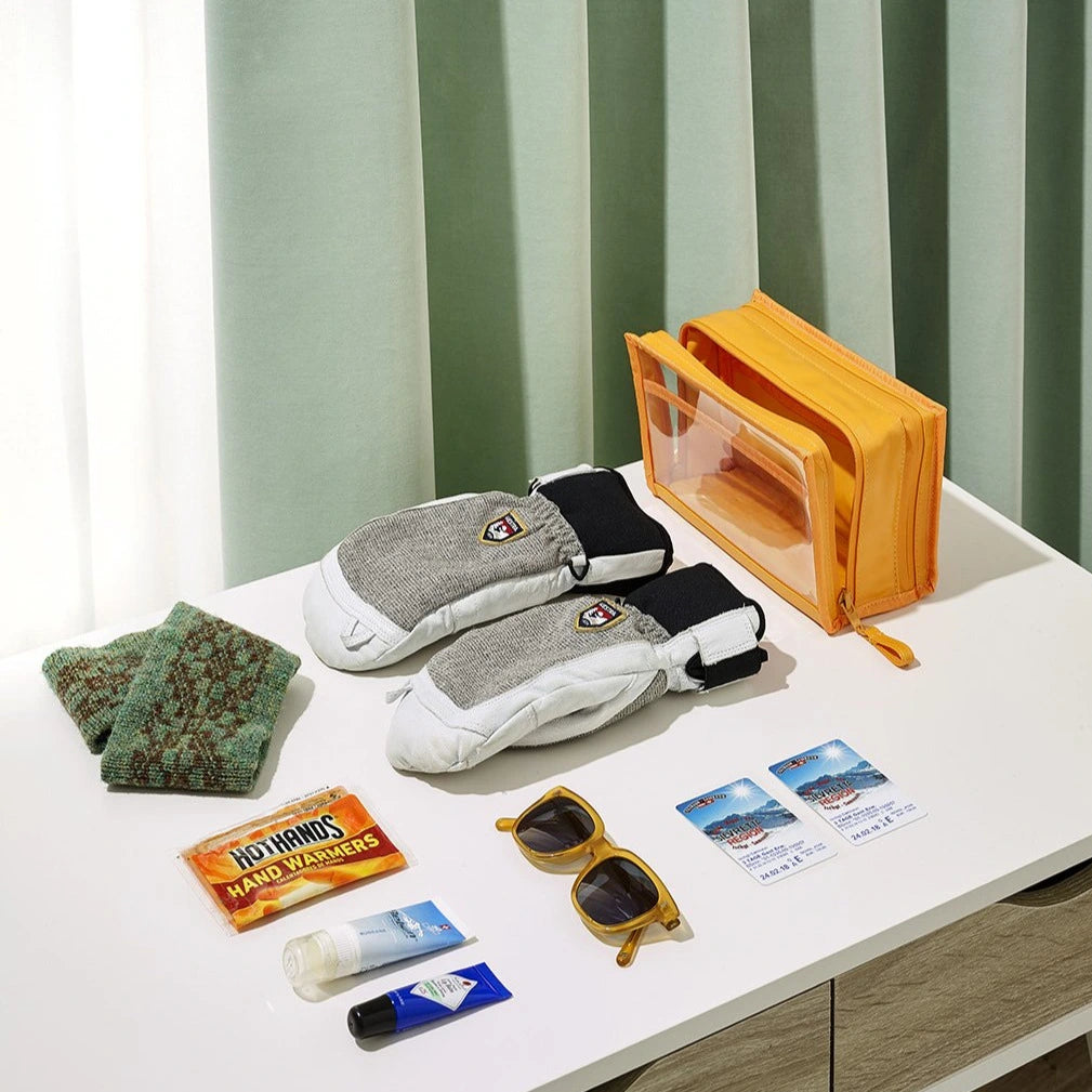 Winter travel essentials flat lay with grey and white ski mittens a vibrant orange pouch yellow sunglasses and Serfaus Fiss Ladis ski tickets