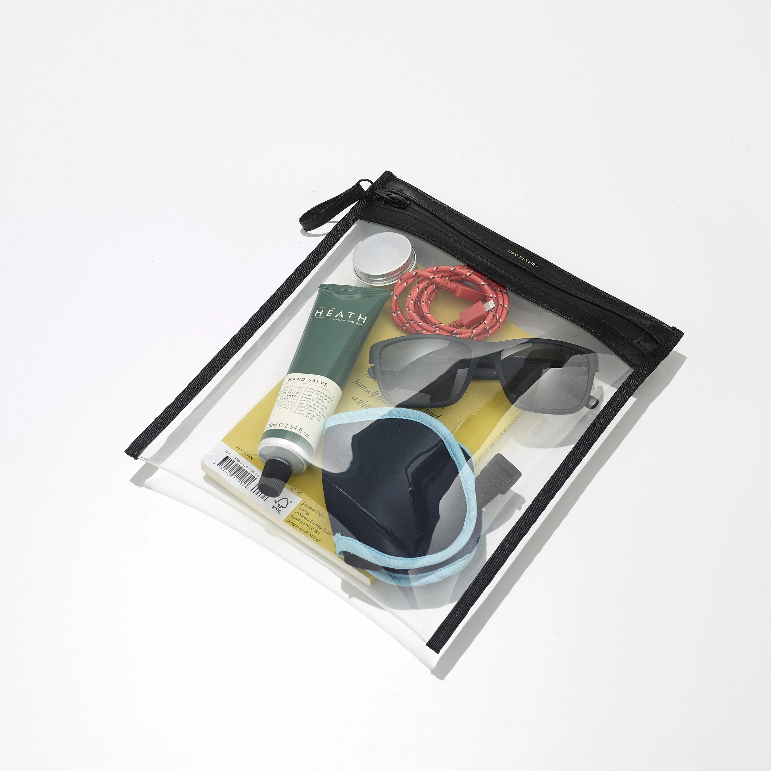 Product photography of a clear zippered travel pouch on a white background filled with a dark green Heath hand salve tube, a coiled coral braided cable, black sunglasses, and a blue and grey sleep mask